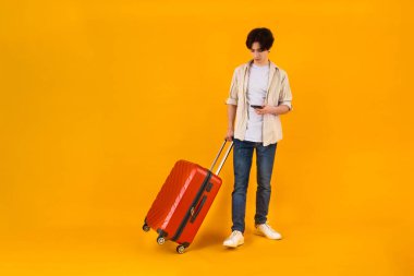 Kırmızı yuvarlanan bir bavulla, sarı bir arka fonda duran günlük kıyafetli bir erkek gezgin..