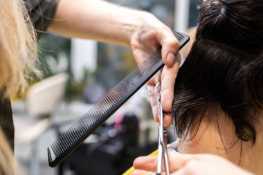 Genç bir adamın saçını tarak ve makasla kesen bir saç stilistinin yakın plan görüntüsü. Görüntü, profesyonel kuaförlükteki hassasiyet ve özeni vurguluyor. Burası modern bir salon..