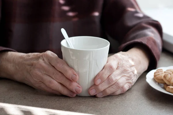 暖かいお茶のカップを保持する老婦人の手 高齢者の手の近くに 自宅でクッキーとお茶を飲み テーブルに座って — ストック写真