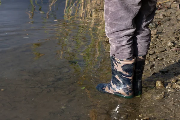 ゴムブーツの子供の足 軍色のレインブーツを着た子供が川岸の水の中に立っている 防水子供の靴 ストックフォト