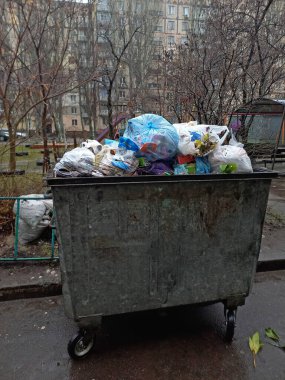 Ev atıklarını toplamak için tekerlekli büyük bir konteynır. Naylon poşetlerdeki çöpler evin bahçesindeki çöp kutusunda duruyor. Dağınık çöp. Açık hava..