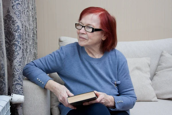 Portrait of elderly woman with closed book in hands, at home, sitting on sofa. Senior woman has finished reading paper book, pondering, looking out window. old woman in reading glasses.