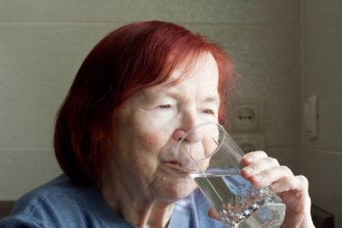 Yaşlı kadın bardaktan maden suyu içiyor. Kırmızı saçlı, mavi kazaklı yaşlı bir kadının yakından çekilmiş portresi susuzluğunu gideriyor. Sağlık ve uzun yaşam için su.