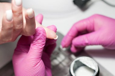 Güzellik salonunda manikür yapma süreci. Pembe eldivenli manikürcü, beyaz tabanı, müşteri elinin tırnaklarına uyguluyor. Profesyonel el bakımı. Sahip ve istemcinin ellerinin kapatılması.