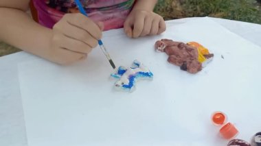 Kız ilkokul öğrencisi boyalarla plastik oyuncak boyuyor. Çocuğun elleri, yakın plan. Yaratıcı yeteneklerin gelişimi, iyi motor beceriler. Yaz. Açık hava.