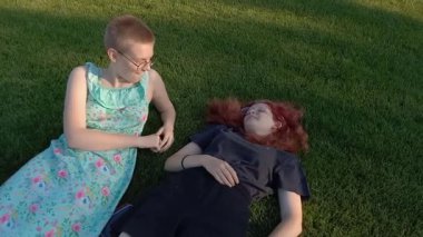 Gözlüklü iki genç kız konuşuyor, dedikodu yapıyor, sohbet ediyor, eğleniyor. Yaz günü şehir parkında yeşil çimlerde yatan kızlar. Modern kızlar. Cihazlar olmadan canlı iletişim.