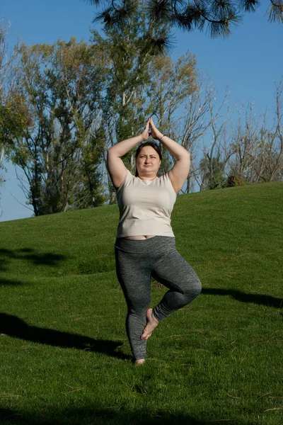 Fitness, outdoor training, weight loss. Fat woman doing exercises in nature. overweight middle-aged woman standing in tree pose, doing yoga in city park. Healthy lifestyle