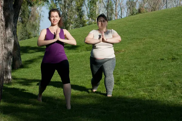 Two middle-aged women practice yoga in city park in warrior pose, standing barefoot on grass. Namaste. Healthy lifestyle, fitness, Pilates, weight loss. overweight woman doing yoga with yoga trainer.