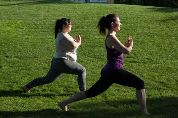 Two middle-aged women practice yoga in city park in warrior pose, standing barefoot on grass. Namaste. Healthy lifestyle, fitness, Pilates, weight loss. overweight woman doing yoga with yoga trainer.