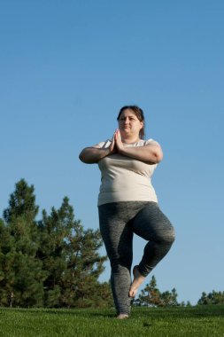 Aşırı kilolu orta yaşlı bir kadın çıplak ayakla yoga yapıyor, tek ayak üzerinde denge egzersizi yapıyor, ağaç duruşu, mavi gökyüzü arka planı. Fitness, yoga, kilo kaybı. Meditasyon, sağlıklı yaşam tarzı.