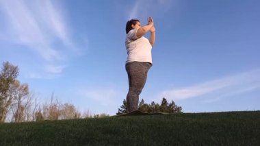 Genç şişman kadın şehir parkında egzersiz yapıyor, mavi gökyüzü geçmişi var. Şişman kadın kilo vermek için spor yapıyor. Aktif açık hava egzersizleri, yoga, güneş selamı. Sağlıklı yaşam tarzı.