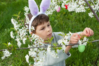 Paskalya yumurtası avı. Anaokulu çocuğu tavşan kulağı takıyor. Bahçede Paskalya yumurtası avında renkli yumurta topluyor. Çiçek açan ağaçların dallarını renkli yumurtalarla süsleyen bir çocuk. Paskalya geleneği