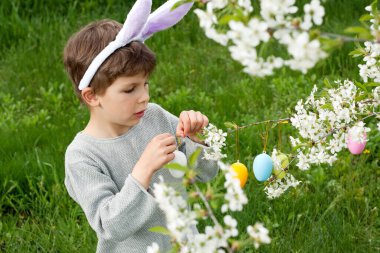 Paskalya yumurtası avı. Anaokulu çocuğu tavşan kulağı takıyor. Bahçede Paskalya yumurtası avında renkli yumurta topluyor. Çiçek açan ağaçların dallarını renkli yumurtalarla süsleyen bir çocuk. Paskalya geleneği