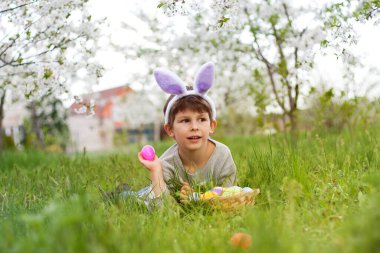 Paskalya yumurtası avı. Bahçedeki Paskalya yumurtası avında yeşil çimenlerin üzerinde yatan tavşan kulaklı sevimli çocuk. Çiçek açan ağaçların arka planına karşı portre. Paskalya geleneği