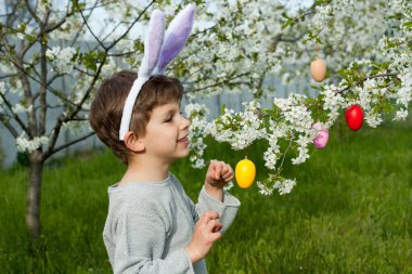 Paskalya yumurtası avı. Tavşan kulağı takan gülümseyen çocuk bahçede Paskalya yumurtası avında renkli yumurtalar buluyor. Çiçek açan ağaçların dallarını renkli yumurtalarla süsleyen bir çocuk. Paskalya geleneği