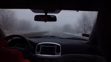 Sisli yol. Araba sis içinde yol boyunca gider, ön camdan bakar. Sınırlı görüş mesafesi, bozuk yol yüzeyi. Bulutlu gri hava. Kışın sisli otoyolda tehlikeli sürüş.