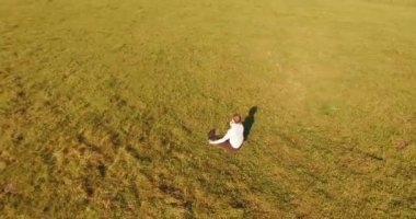 Havadan bakış açısı. Yeşil çimlerde oturan ve kırsal alanda not defteri olan bir adamın etrafında alçak yörünge uçuşları. Günlük giysi ve zen meditasyon pozu. Güneş ışınları. Radikal hareket.