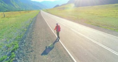 Hava 4K UHD görüntüsü. Asfalt yolda yürüyen otostopçu turist üzerinde alçak irtifa uçuşu. Büyük bir kırsal vadi ve yaz günü güneşli bir çayır. Sırt çantalı gezgin..