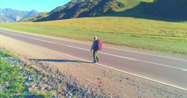Hava 4K UHD görüntüsü. Asfalt yolda yürüyen otostopçu turist üzerinde alçak irtifa uçuşu. Büyük bir kırsal vadi ve yaz günü güneşli bir çayır. Sırt çantalı gezgin..