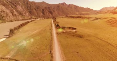 Hava UHD 4K: güneşli yaz sabahında kırsal dağ yolu ve çayır üzerinde havadan uçuş. Ufukta yeşil ağaçlar ve güneş ışınları var. Özgür kırsal asfalt karayolu ve nehir.