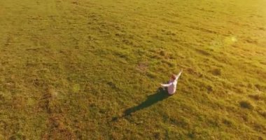 Havadan bakış açısı. Yeşil çimlerde oturan ve kırsal alanda not defteri olan bir adamın etrafında alçak yörünge uçuşları. Gündelik kıyafet ve Zen meditasyonu. Eller havada. Güneş ışınları. Radikal hareket.
