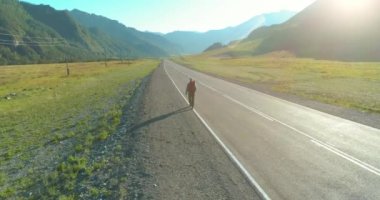 Hava 4K UHD görüntüsü. Asfalt yolda yürüyen otostopçu turist üzerinde alçak irtifa uçuşu. Büyük bir kırsal vadi ve yaz günü güneşli bir çayır. Sırt çantalı gezgin..
