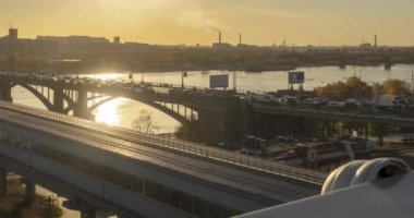 Şehir hayatının 4K UHD zaman çizelgesi. Güneş ışınları, mavi gökyüzü, ufukta nehir ve köprü. Yaz günbatımında trafik sıkışık. Motorlu kaydırma hareketi