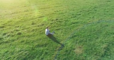 Havadan bakış açısı. Yeşil çimlerde oturan ve kırsal alanda not defteri olan bir adamın etrafında alçak yörünge uçuşları. Günlük giysi ve zen meditasyon pozu. Güneş ışınları. Radikal hareket.