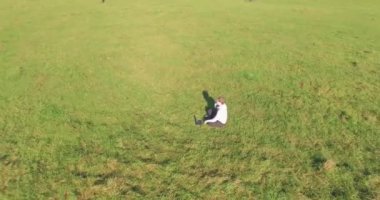 Havadan bakış açısı. Yeşil çimlerin üzerinde oturan, sarı kırsal alanda not defteri olan ve telefonla konuşan bir adam. Günlük kıyafet. Güneş ışınları. Radikal hareket.