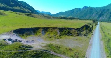 Hava UHD 4K: güneşli yaz sabahında kırsal dağ yolu ve çayır üzerinde havadan uçuş. Ufukta yeşil ağaçlar ve güneş ışınları var. Özgür kırsal asfalt karayolu ve nehir.