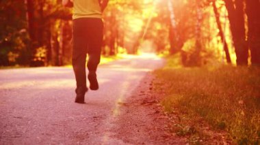 Yeni asfalt yolda koşan FHD sporcusu. Şehir parkında gün doğumu ya da gün batımı. Yeşil ağaç ormanı ve ufukta güneş ışığı.
