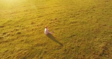 Havadan bakış açısı. Yeşil çimlerde oturan ve kırsal alanda not defteri olan bir adamın etrafında alçak yörünge uçuşları. Günlük giysi ve zen meditasyon pozu. Güneş ışınları. Radikal hareket.