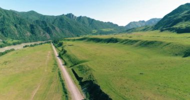 Hava UHD 4K: güneşli yaz sabahında kırsal dağ yolu ve çayır üzerinde havadan uçuş. Ufukta yeşil ağaçlar ve güneş ışınları var. Özgür kırsal asfalt karayolu ve nehir.