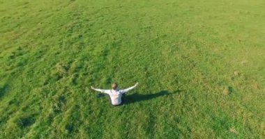 Havadan bakış açısı. Yeşil çimlerde oturan ve kırsal alanda not defteri olan bir adamın etrafında alçak yörünge uçuşları. Gündelik kıyafet ve Zen meditasyonu. Eller havada. Güneş ışınları. Radikal hareket.