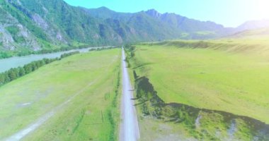 Hava UHD 4K: güneşli yaz sabahında kırsal dağ yolu ve çayır üzerinde havadan uçuş. Ufukta yeşil ağaçlar ve güneş ışınları var. Özgür kırsal asfalt karayolu ve nehir.