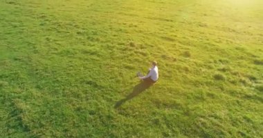 Havadan bakış açısı. Yeşil çimlerde oturan ve kırsal alanda not defteri olan bir adamın etrafında alçak yörünge uçuşları. Günlük giysi ve zen meditasyon pozu. Güneş ışınları. Radikal hareket.