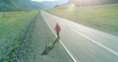 Hava 4K UHD görüntüsü. Asfalt yolda yürüyen otostopçu turist üzerinde alçak irtifa uçuşu. Büyük bir kırsal vadi ve yaz günü güneşli bir çayır. Sırt çantalı gezgin..