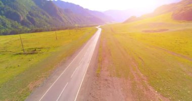 Hava UHD 4K görüntüsü. Güneşli yaz sabahında, dağ asfaltı yolu ve çayır üzerinde alçak uçuş. Yeşil ağaçların yanında. Güneş ışınlarıyla hızlı yatay hareket.