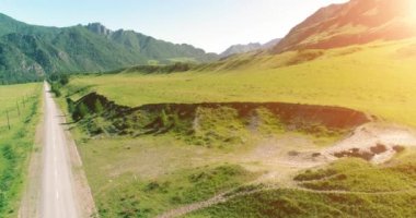 Hava UHD 4K: güneşli yaz sabahında kırsal dağ yolu ve çayır üzerinde havadan uçuş. Ufukta yeşil ağaçlar ve güneş ışınları var. Özgür kırsal asfalt karayolu ve nehir.