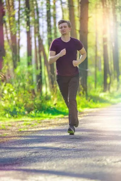 A man wearing a hat is jogging along an asphalt road surrounded by grass and trees in the woods for leisure and recreation