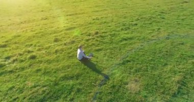 Havadan bakış açısı. Yeşil çimlerde oturan ve kırsal alanda not defteri olan bir adamın etrafında alçak yörünge uçuşları. Günlük giysi ve zen meditasyon pozu. Güneş ışınları. Radikal hareket.