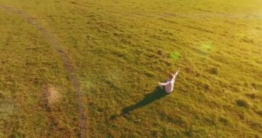 Havadan bakış açısı. Yeşil çimlerde oturan ve kırsal alanda not defteri olan bir adamın etrafında alçak yörünge uçuşları. Gündelik kıyafet ve Zen meditasyonu. Eller havada. Güneş ışınları. Radikal hareket.