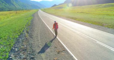 Hava 4K UHD görüntüsü. Asfalt yolda yürüyen otostopçu turist üzerinde alçak irtifa uçuşu. Büyük bir kırsal vadi ve yaz günü güneşli bir çayır. Sırt çantalı gezgin..