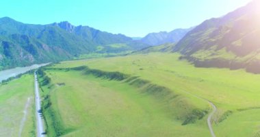 Hava UHD 4K: güneşli yaz sabahında kırsal dağ yolu ve çayır üzerinde havadan uçuş. Ufukta yeşil ağaçlar ve güneş ışınları var. Özgür kırsal asfalt karayolu ve nehir.