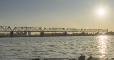 Şehir nehir kıyısının 4K UHD zaman çizelgesi. Güneş ışınları, mavi gökyüzü ve ufukta demiryolu köprüsü. Düz küçük dalgalar, su ve motorlu kaydırak hareketleriyle yaz günbatımı zamanı