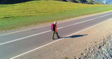 Hava 4K UHD görüntüsü. Asfalt yolda yürüyen otostopçu turist üzerinde alçak irtifa uçuşu. Büyük bir kırsal vadi ve yaz günü güneşli bir çayır. Sırt çantalı gezgin..