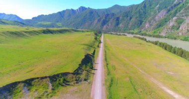 Hava UHD 4K: güneşli yaz sabahında kırsal dağ yolu ve çayır üzerinde havadan uçuş. Ufukta yeşil ağaçlar ve güneş ışınları var. Özgür kırsal asfalt karayolu ve nehir.