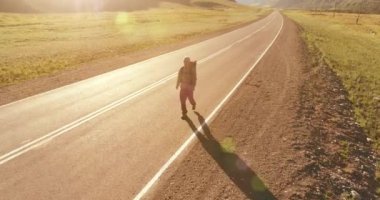 Hava 4K UHD görüntüsü. Asfalt yolda yürüyen otostopçu turist üzerinde alçak irtifa uçuşu. Büyük bir kırsal vadi ve yaz günü güneşli bir çayır. Sırt çantalı gezgin..