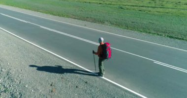 Hava 4K UHD görüntüsü. Asfalt yolda yürüyen otostopçu turist üzerinde alçak irtifa uçuşu. Büyük bir kırsal vadi ve yaz günü güneşli bir çayır. Sırt çantalı gezgin..