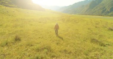 Hava 4K UHD görüntüsü. Yeşil dağ tarlasında yürüyen genç turist üzerinde alçak irtifa uçuşu. Büyük bir kırsal vadi ve yaz günü güneşli bir çayır. Sırt çantalı gezgin..